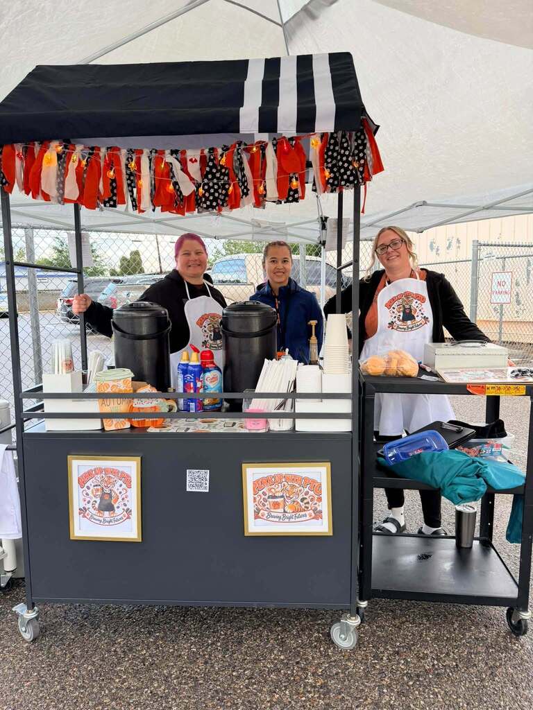 Three staff members smile behind a decorated “Perk Up with PTO” coffee and snack cart featuring orange, black, and white decorations.