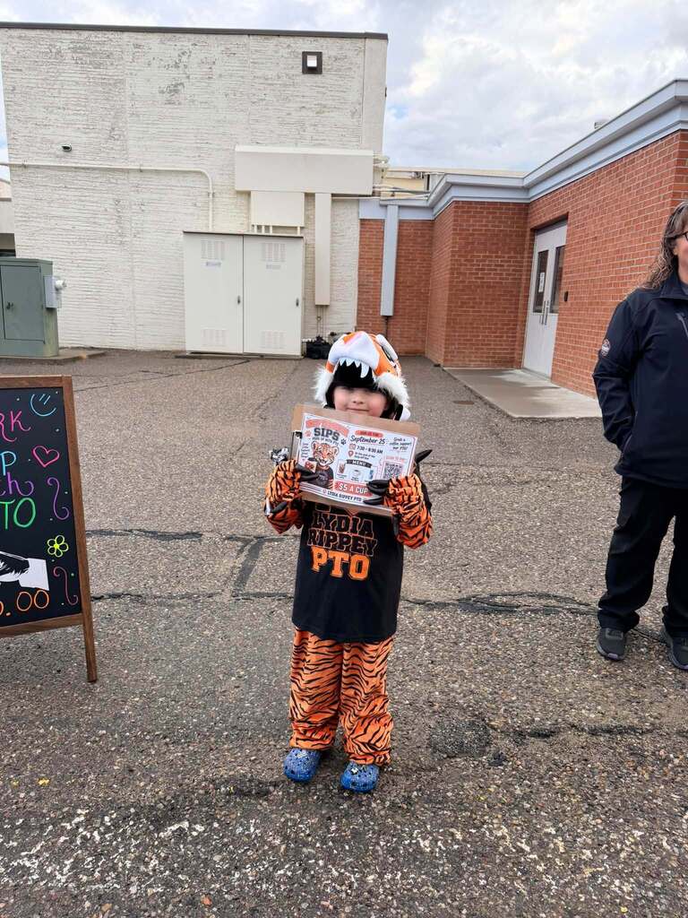 A student dressed in an orange and black tiger costume holds a flyer for the “Perk Up with PTO” event outside a school building.