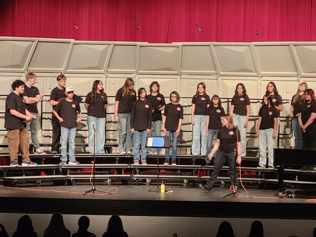 Koogler Middle School choir students stand on risers on stage during a choir performance, with a student and microphone in the foreground.