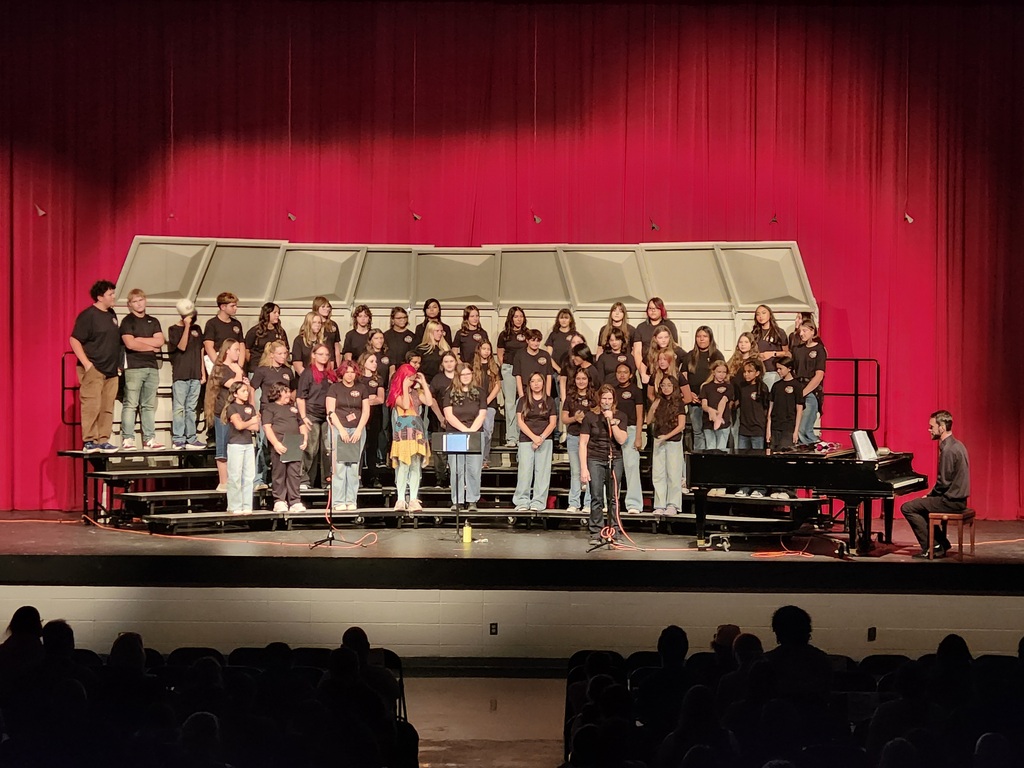 Koogler Middle School choir students stand on risers on stage during a choir performance, with a student and microphone in the foreground.