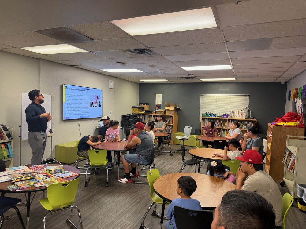 A presenter stands at the front of a room at Esperanza Elementary School, speaking to families during an ASU WeGrad educational session. A large screen displays a presentation in Spanish. Parents, guardians, and children sit together at round tables, listening and participating in the discussion. Bookshelves, colorful chairs, and educational materials create a welcoming learning environment.