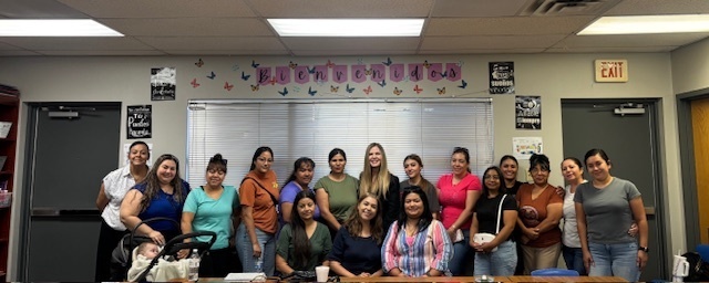 A group photo of parents and a facilitator standing and sitting together inside the same classroom. Everyone is smiling at the camera. The wall behind them has a large banner that reads “Bienvenidos” with butterfly decorations.