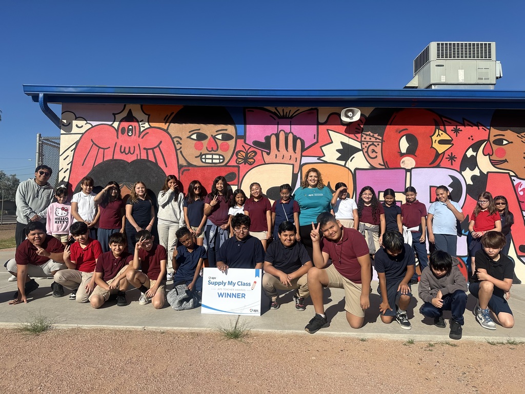 A large group of students pose outside with their teacher and an APS representative in front of a brightly colored mural featuring cartoon-style faces and designs. One student in the front row holds the same sign that reads, “APS Supply My Class – APS Teacher Awards – Winner.” The group smiles and makes peace signs while standing under a clear blue sky.