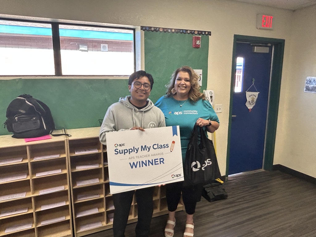 A teacher stands indoors holding a large sign that reads, “APS Supply My Class – APS Teacher Awards – Winner.” Next to him is a woman smiling, wearing a teal APS Community Connectors shirt, and holding a black APS tote bag. They are in a classroom with cubby shelves behind them and a blue door to the right.