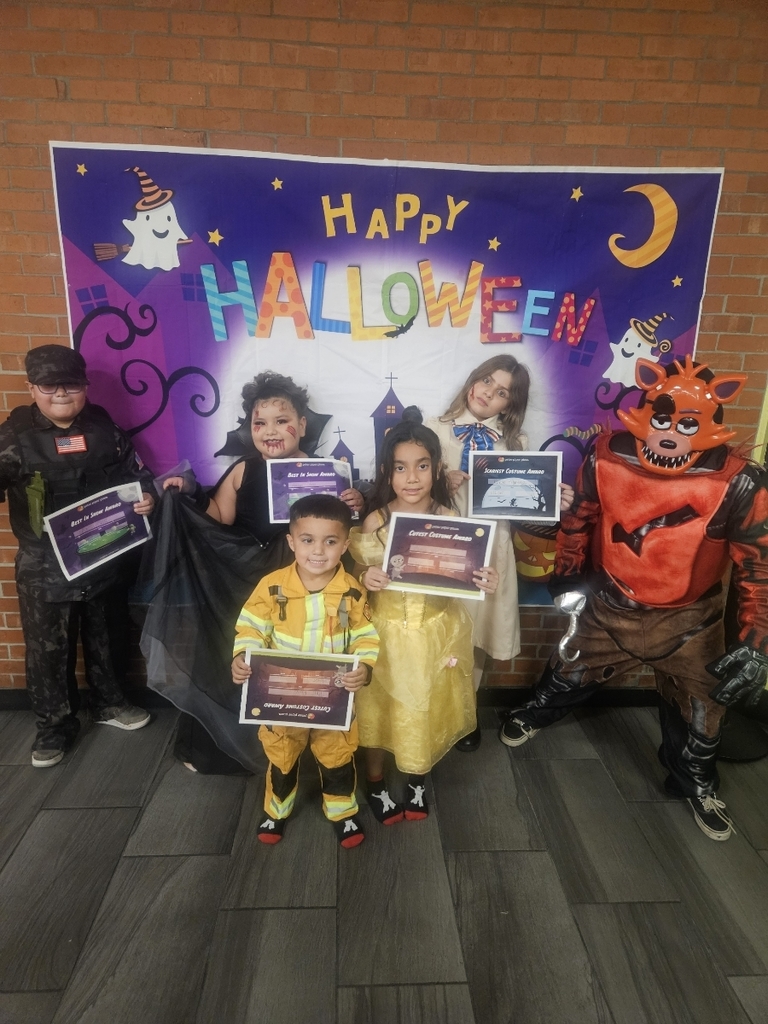 Five children posing together in costume in front of a large "Happy Halloween" backdrop. The children are holding various awards or certificates for the costume contest. Costumes include: a dark, tactical military outfit, a vampire, a small firefighter, a Belle-like princess in a yellow dress, a classic princess with a bow, and a Foxy character from Five Nights at Freddy's.