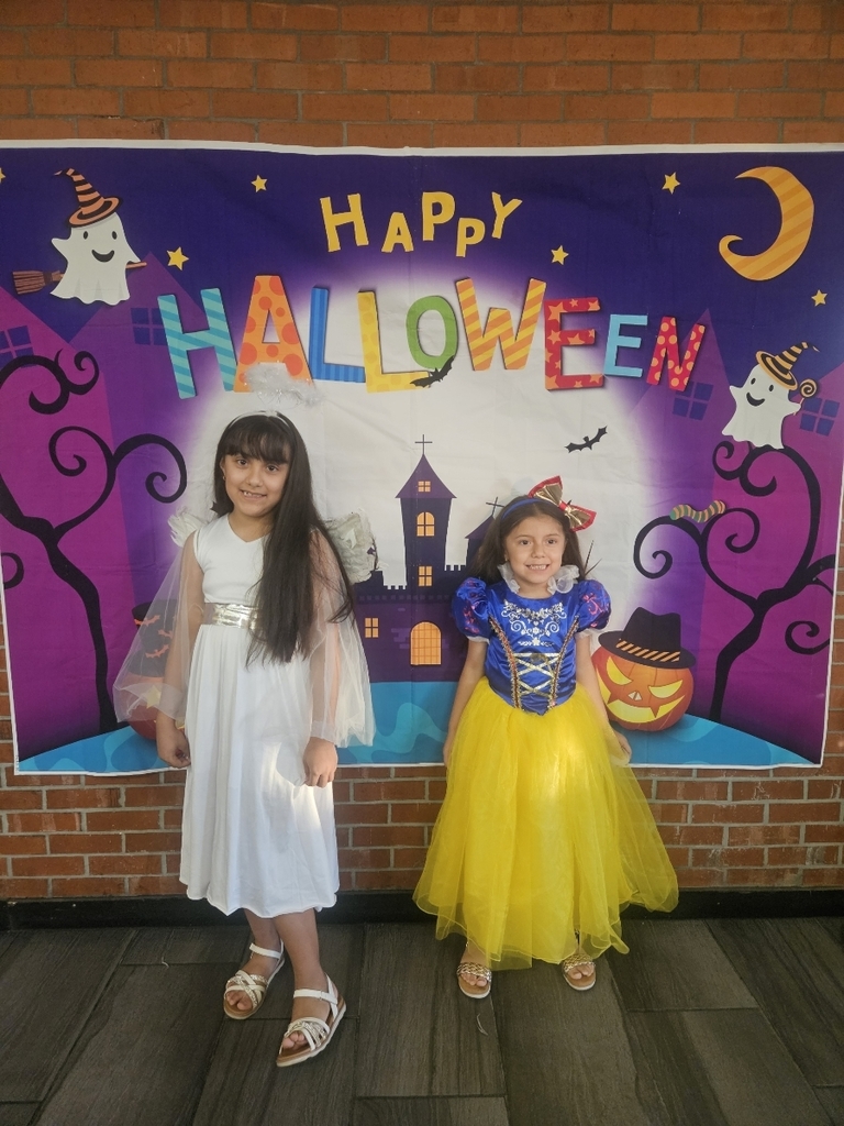 Two young girls smiling in front of a Halloween backdrop featuring a castle, ghosts, and the text "Happy Halloween." The girl on the left is dressed as an angel in a long white dress with sheer white wings. The girl on the right is dressed as Snow White in a blue, red, and yellow princess gown. They are standing on a dark floor in front of a brick wall.