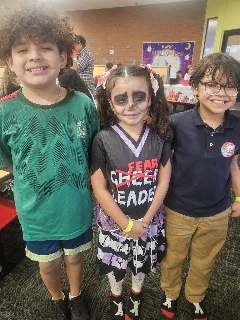 Three children posing and smiling for the camera in a brightly lit indoor space. The boy on the left is wearing a green Mexico soccer jersey. The girl in the center has face paint to look like a zombie or skeleton and is wearing a black and purple "Fear Cheerleader" costume. The boy on the right is wearing a dark blue polo shirt and glasses.
