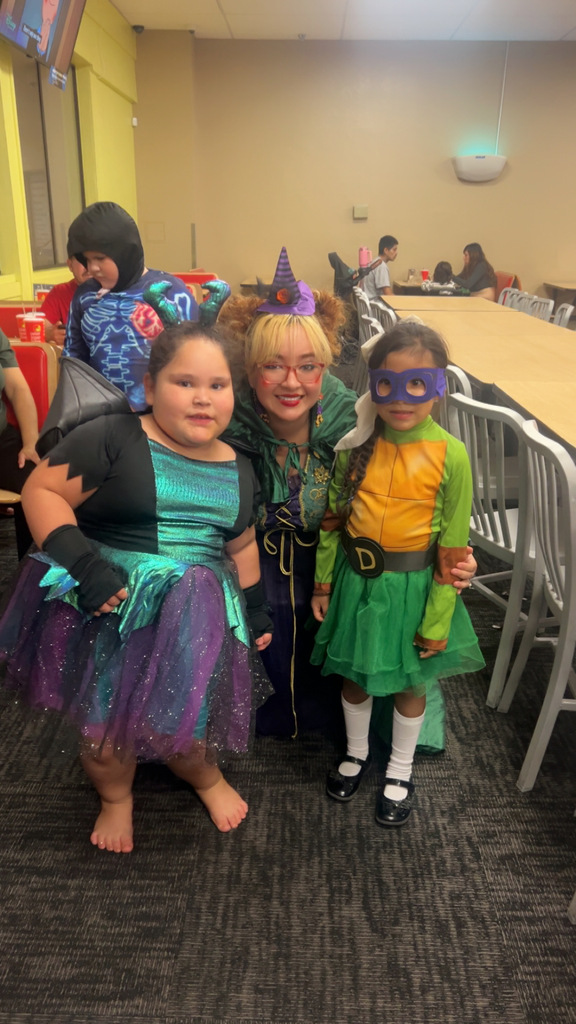 A woman dressed as a witch kneels between two girls who are in costume. The girl on the left is wearing a sparkly black and purple fairy or witch costume. The girl on the right is wearing a green and brown Donatello-themed Teenage Mutant Ninja Turtles costume with a purple mask. They are indoors on a dark carpet.
