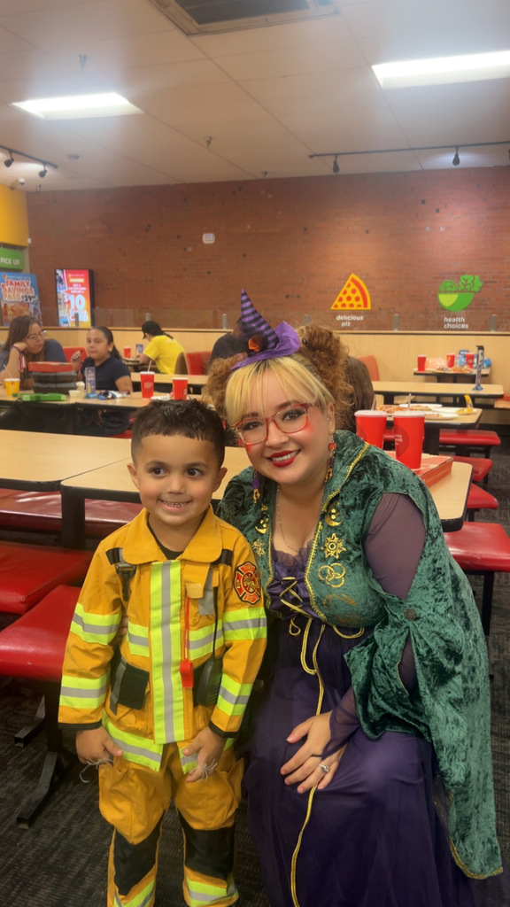A woman dressed as a witch in a green velvet robe and a purple hat kneels next to a young boy. The boy is smiling and dressed as a firefighter in a yellow coat and pants with reflective stripes. They are inside a Peter Piper Pizza restaurant with red booths and tables visible in the background.