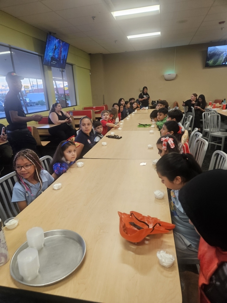 A long-shot of a group of children seated along a long, rectangular table inside the restaurant. They appear to be engaged in an activity or waiting for food. A few adults are visible on the left and at the far end of the table. A tray with plastic cups and an orange plastic item sits on the table in the foreground.