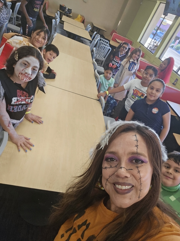 A group selfie showing a mix of children and adults seated around a long, light-brown table at an indoor event space. A woman in the foreground is smiling at the camera and has white stitches painted onto her face and a festive garland in her hair. Several children around the table are wearing various costumes and face paint, including a zombie cheerleader and a boy in a green Minecraft-themed shirt.