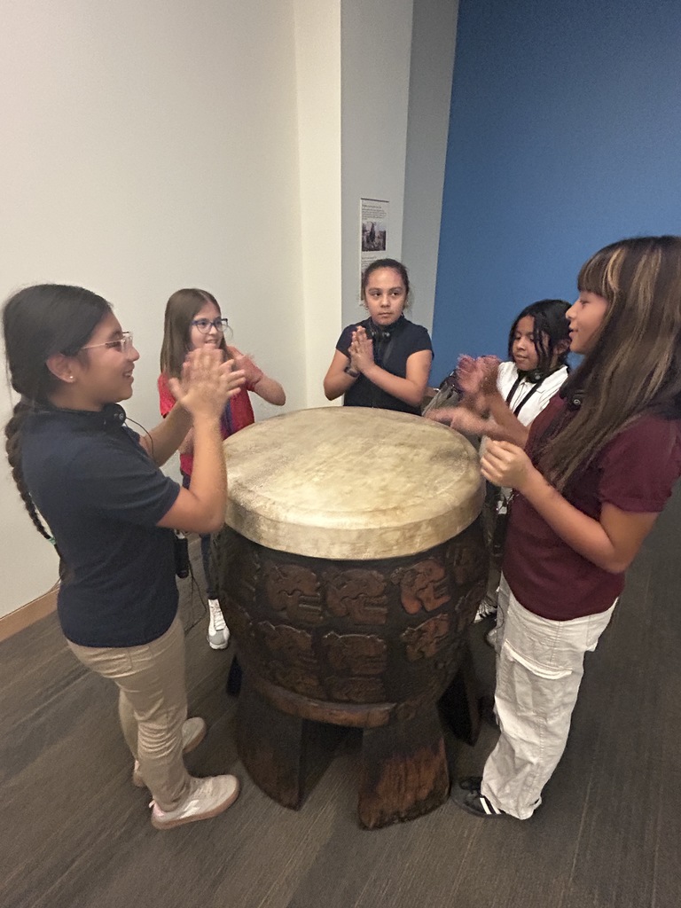 A close-up photo of five students standing around a large, ancient-looking, carved wooden drum with a wide, light-colored goatskin head. The students are smiling and seem to be clapping or patting the drum's head.