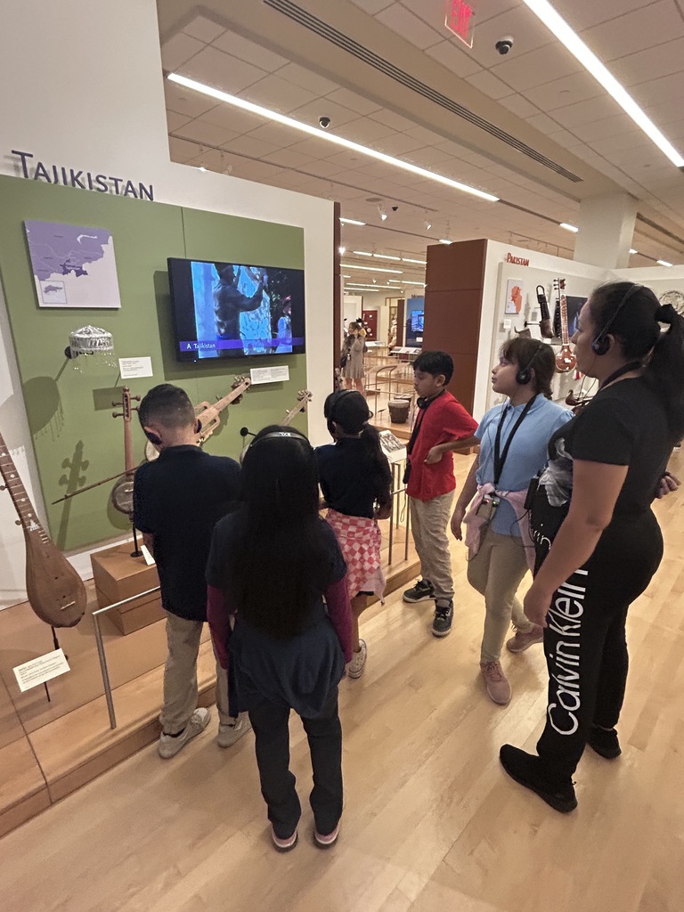 Five students stand in front of an exhibit labeled "Tajikistan" and "Pakistan." They are looking at a wall-mounted video screen that displays a musician. Traditional string and plucked instruments hang on the walls around the screen.