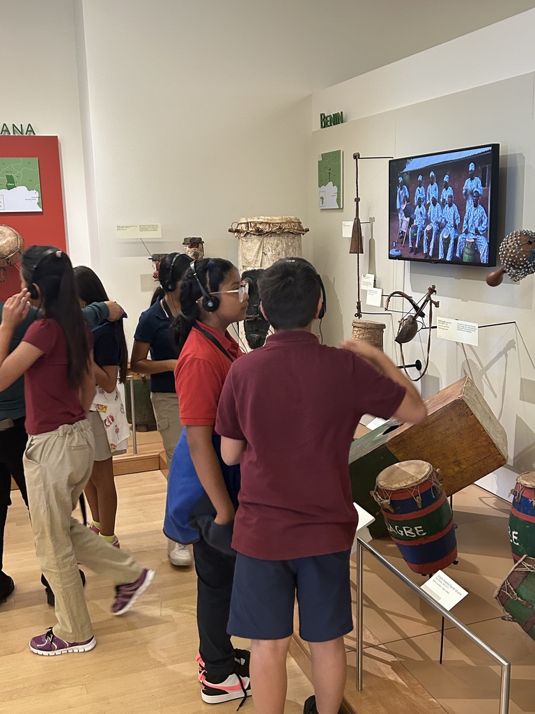 A photo of three students wearing headphones, standing in front of an exhibit wall labeled "Benin." The exhibit features various West African drums and instruments, including a large wooden drum and a screen showing a video of musicians