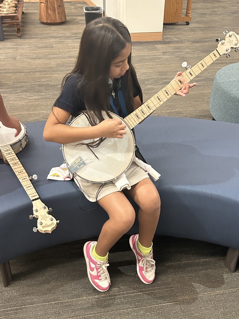 A close-up, seated photo of a young girl wearing headphones and concentrating while she plays a banjo. Another banjo is resting on the blue upholstered bench next to her.
