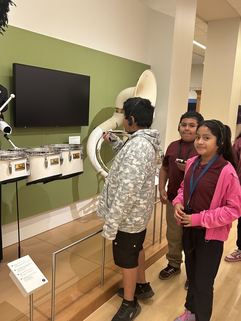 Three students stand next to a large brass tuba displayed behind a railing and a set of white drums. The students are wearing headphones and smiling, one is looking directly at the camera.