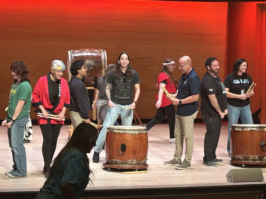 A group of nine adults, some of whom are holding drumsticks, standing on a stage with several large Japanese Taiko drums. The stage has a red background wall, and the adults appear to be part of a demonstration or performance.