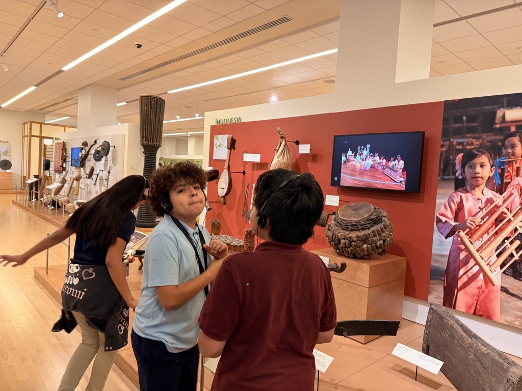 Two students, wearing headphones, stand in conversation in front of a red exhibit wall labeled "Indonesia." The display features several string instruments, a woven drum, and a large photo of a young girl holding an Angklung (a bamboo instrument).