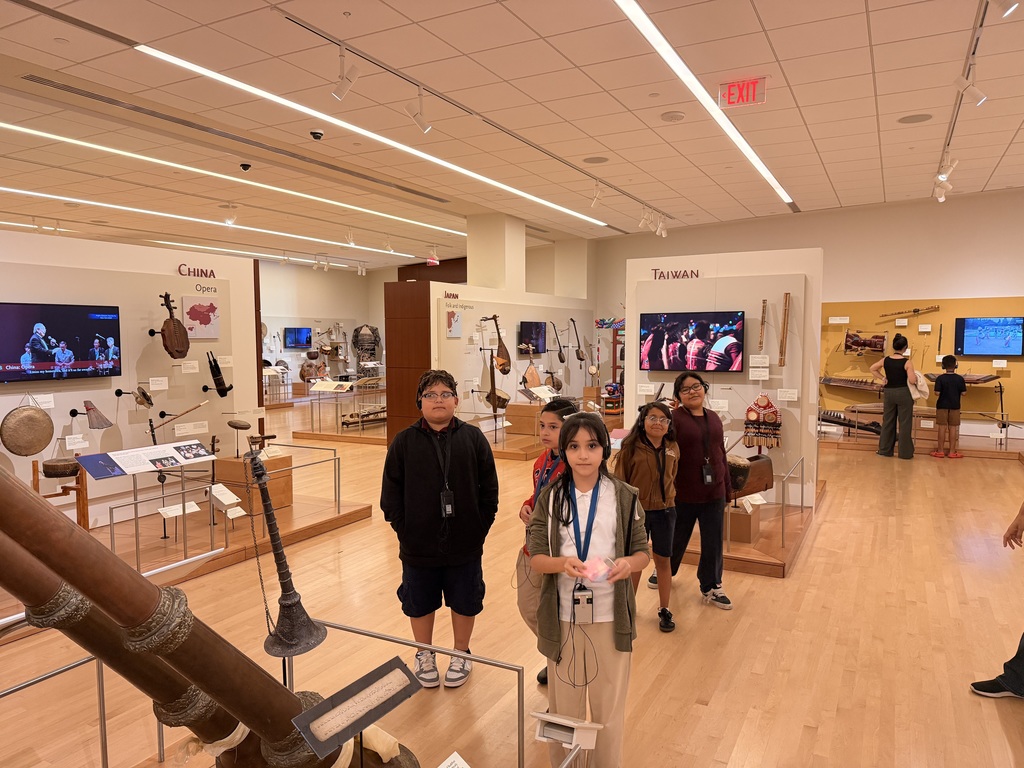Five students are standing and looking at a wide, brightly lit gallery showcasing instruments from China and Taiwan. The walls are filled with various traditional instruments, and video screens display musicians.