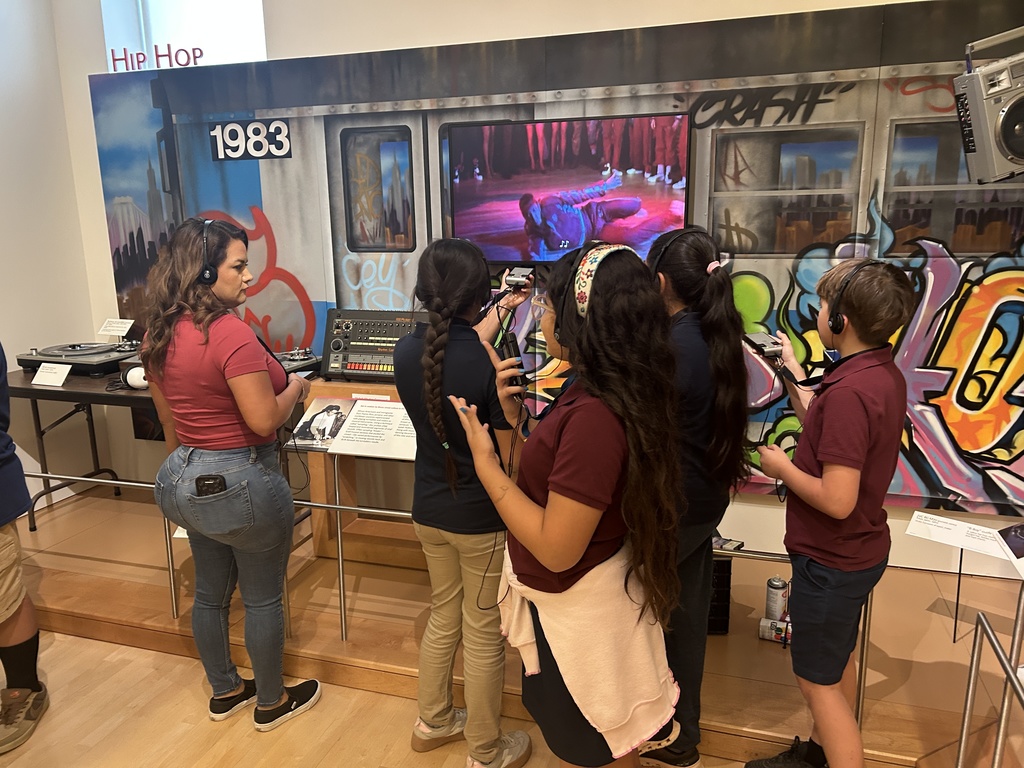 A group of five students and one adult standing in front of an exhibit on the history of Hip-Hop. The backdrop is a mural depicting a subway car with "1983" painted on it and graffiti on the wall. The students are wearing headphones, looking intently at a video screen, and interacting with a small DJ mixing board on a table.
