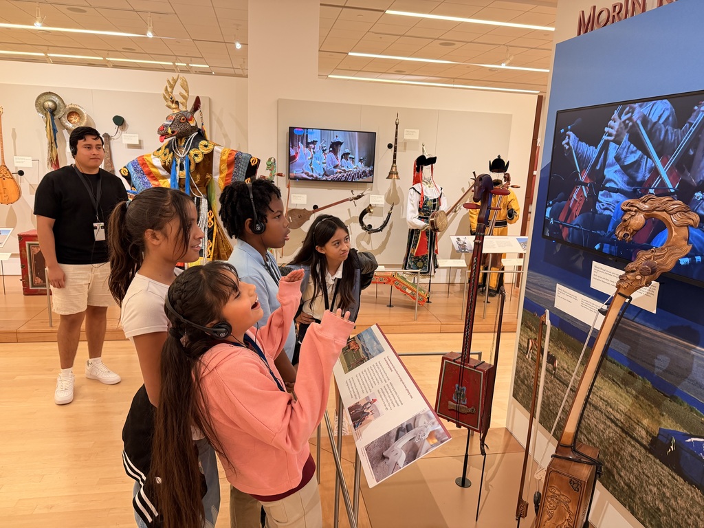 Five students and a male chaperone stand in the gallery viewing an exhibit on Mongolian music. They are looking at a display panel that features a large photo of musicians, a detailed carved instrument head (likely a morin khuur, or horse-head fiddle), and a book with text and images. The students are wearing headphones.