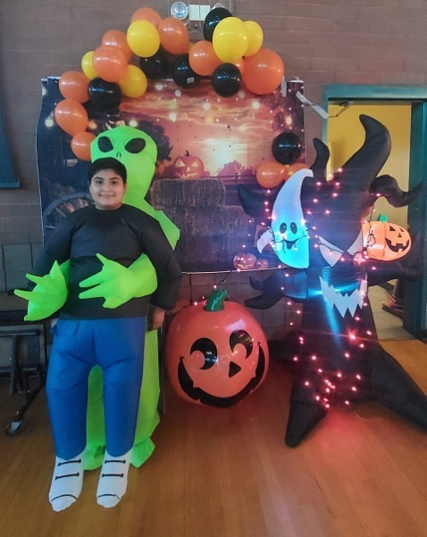 Young boy standing in front of a fall Halloween decorated photo booth with balloons and a jack-o-lantern. The boy is wearing an inflatable costume that looks like a green alien is holding him.