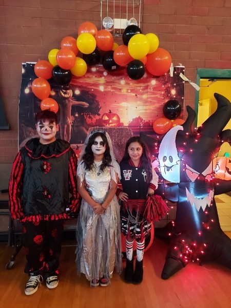 3 students pose in front of a fall/halloween decorated backdrop. One student is dressed as a clown, one as the corpse-bride, and the last is dressed as a cheerleader