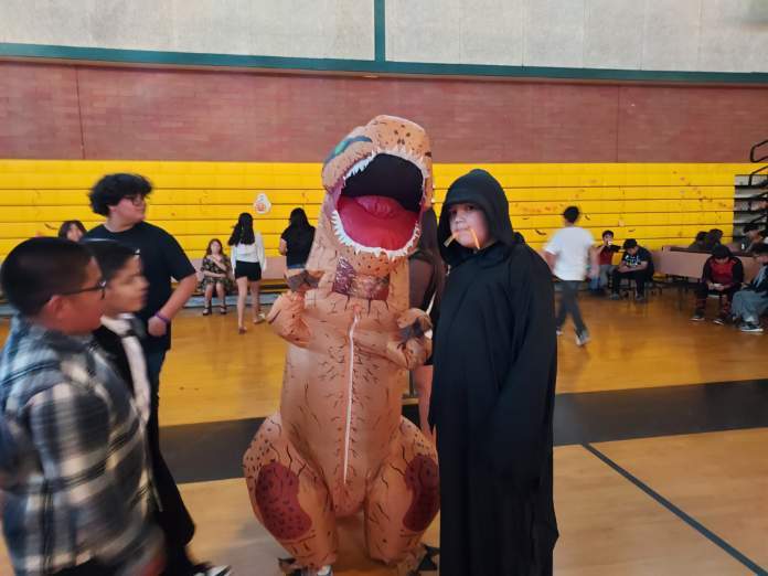 Two boys. One dressed up in an orange inflatable t-rex costume; the other in a black cloak with a hood