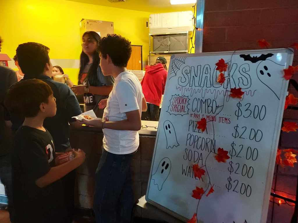 Students stand in line for snacks. There is a sign stating what is served and for how much. The sign has ghosts and spiderwebs drawn on it.