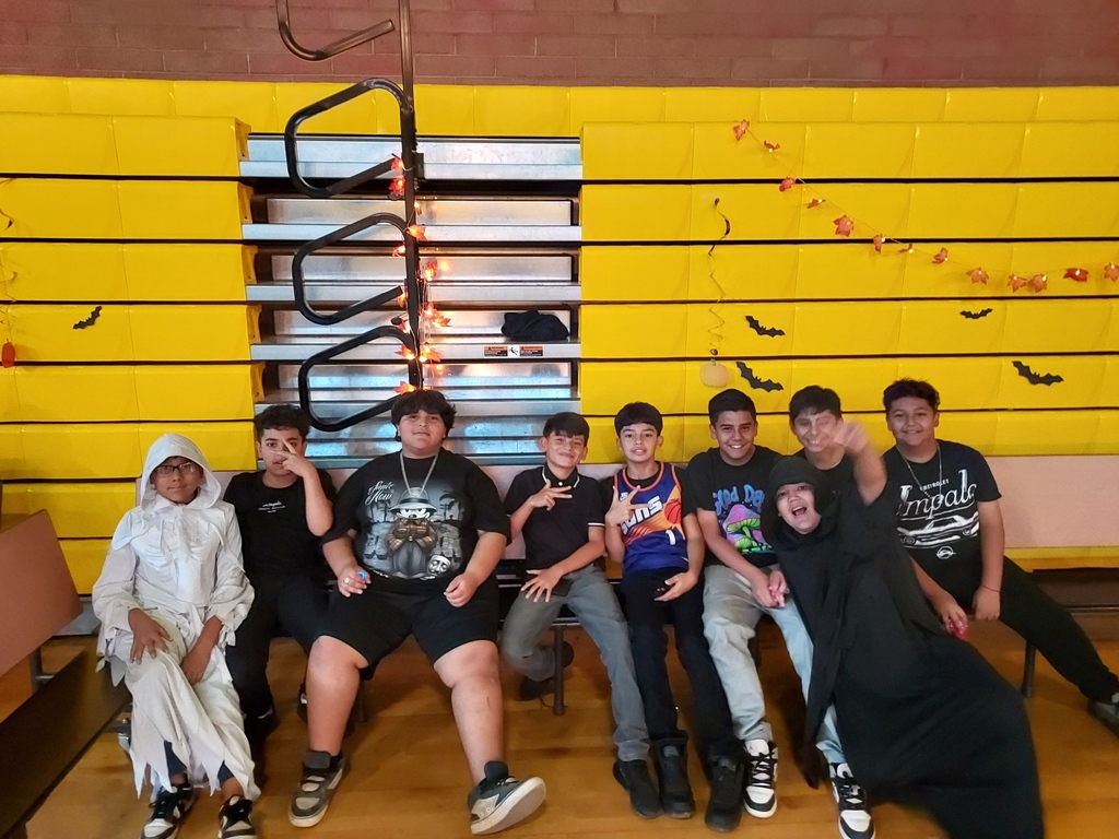 9 boy students sit on a bench. All wearing different costumes. The background has paper bats and other fall decorations