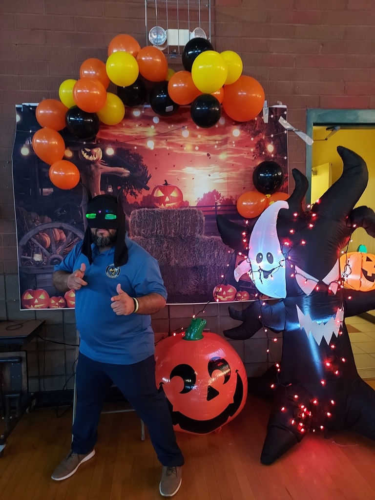 A teacher wearing a Batman mask stands in front of a fall/Halloween backdrop