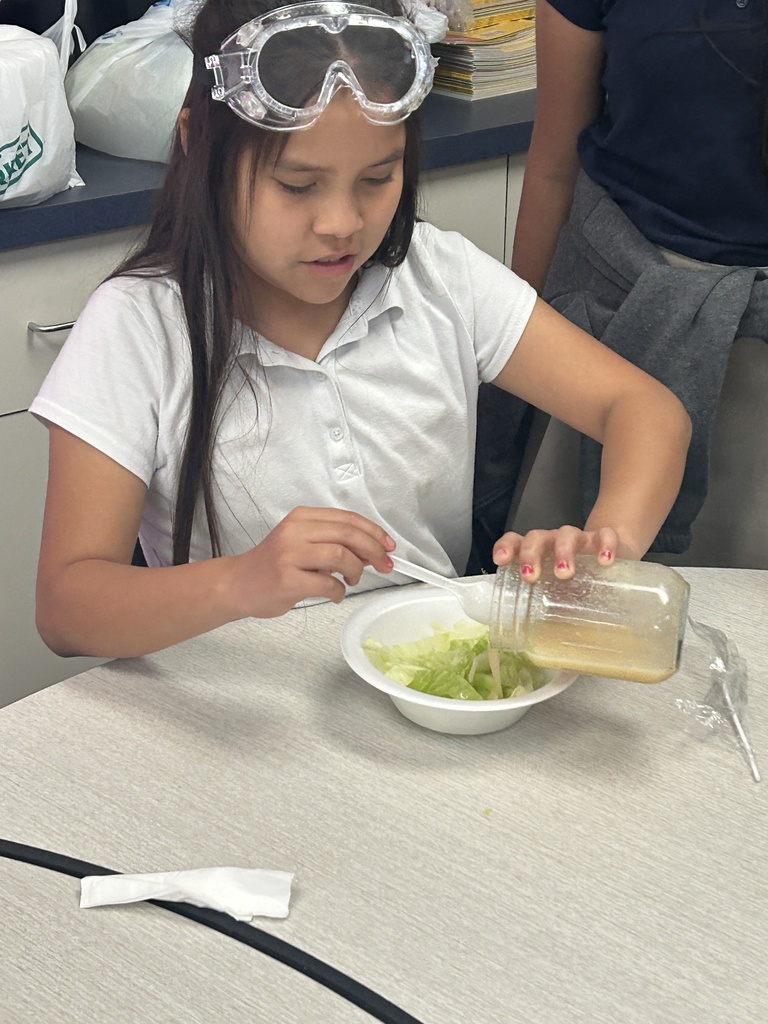A 5th-grade girl, wearing safety goggles pushed up on her head, smiles slightly as she pours her finished homemade salad dressing from a glass jar onto a small white bowl of shredded lettuce. This photo captures the final step of tasting their scientific creation.