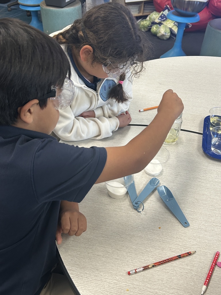 A 5th-grade boy wearing safety goggles uses a small measuring spoon to add a white solid ingredient to a glass jar. A girl wearing goggles is leaning on the table next to him, observing the process. Clear cups with ingredients are visible on a blue tray in the background.