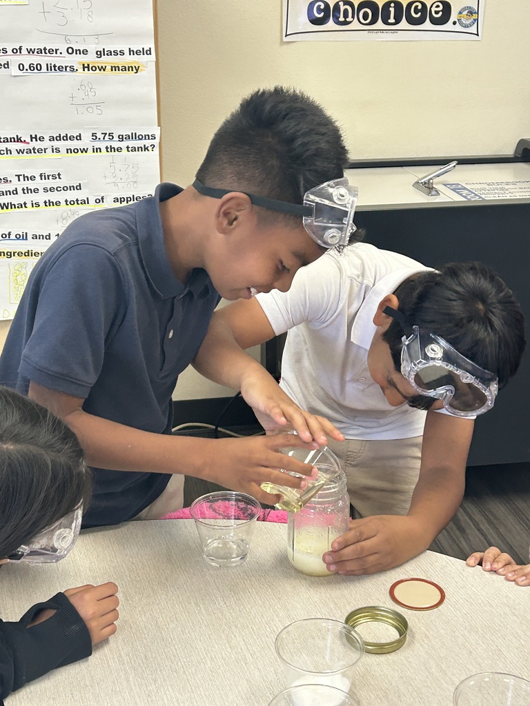 Two 5th-grade boys, both wearing safety goggles, are collaborating to make their salad dressing. The boy on the left is carefully pouring a yellow liquid (likely oil) from a beaker into a glass jar held by the boy on the right, which contains a white liquid. The background shows part of a math anchor chart on the wall.