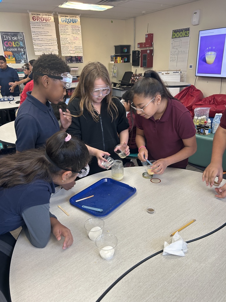 Four 5th-grade students, wearing safety goggles, are gathered around a circular table to mix ingredients for their salad dressing. One student pours a liquid from a small clear container into a larger jar held by another student, while a third student stirs a substance in a small cup. A clear bowl containing milk or a white liquid sits on the table, and they are using a blue tray for some of their materials.