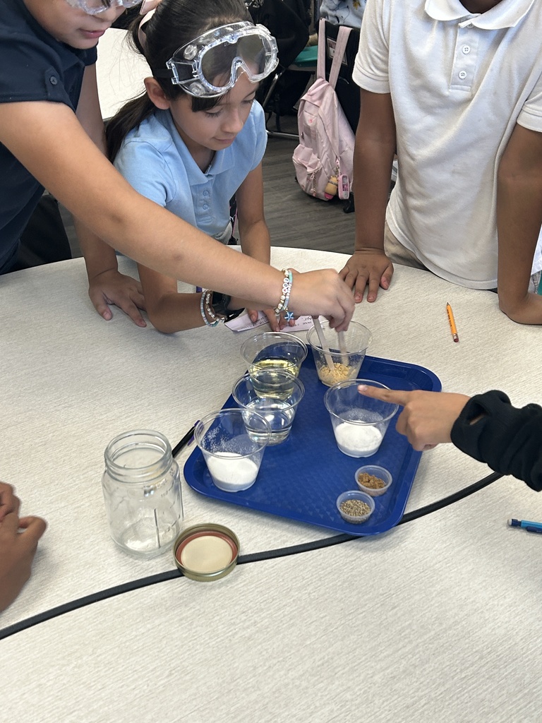 A group of students, one wearing safety goggles, stands around a table observing and mixing ingredients. On a blue tray are several small cups containing measured ingredients for the salad dressing, including oil, water, and white granulated solids (salt or sugar). One student's hand points toward the ingredients, while another student stirs a cup. A glass jar and its lid are visible to the side.