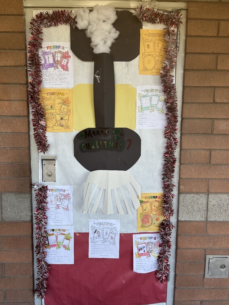 A door decorated to look like a fireplace or chimney. A large black shape resembling a stovepipe or chimney rises up the center with white, cotton-like material at the top to simulate smoke. A red skirt-like base covers the bottom of the door. Various pages with handwritten "Thankful" notes and drawings are taped around the central chimney structure, and red tinsel frames the door.