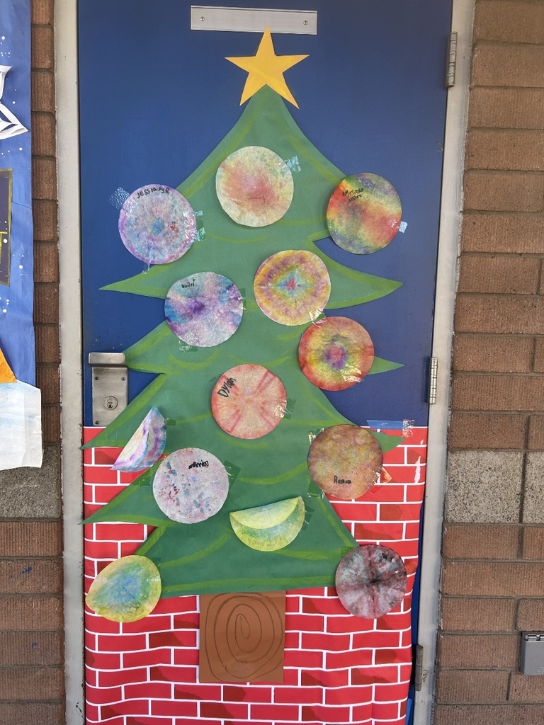 A door decorated with a large green paper Christmas tree shape over a red and white brick-patterned backdrop (to resemble a fireplace). The ornaments on the tree are made from colorful, tie-dye style coffee filters, with some names written on them. A yellow star tops the tree.