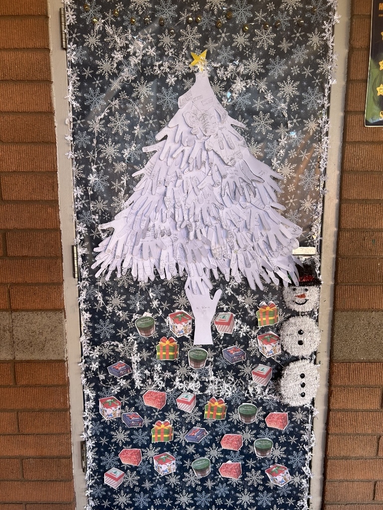 A door covered in a dark blue/black paper with a white snowflake pattern. In the center, a Christmas tree is constructed entirely from cut-out white paper handprints, topped with a yellow star. Beneath the tree are multiple small, colorful gift and cup cutouts, and a snowman figure made from three white fuzzy pom-poms is on the right side.