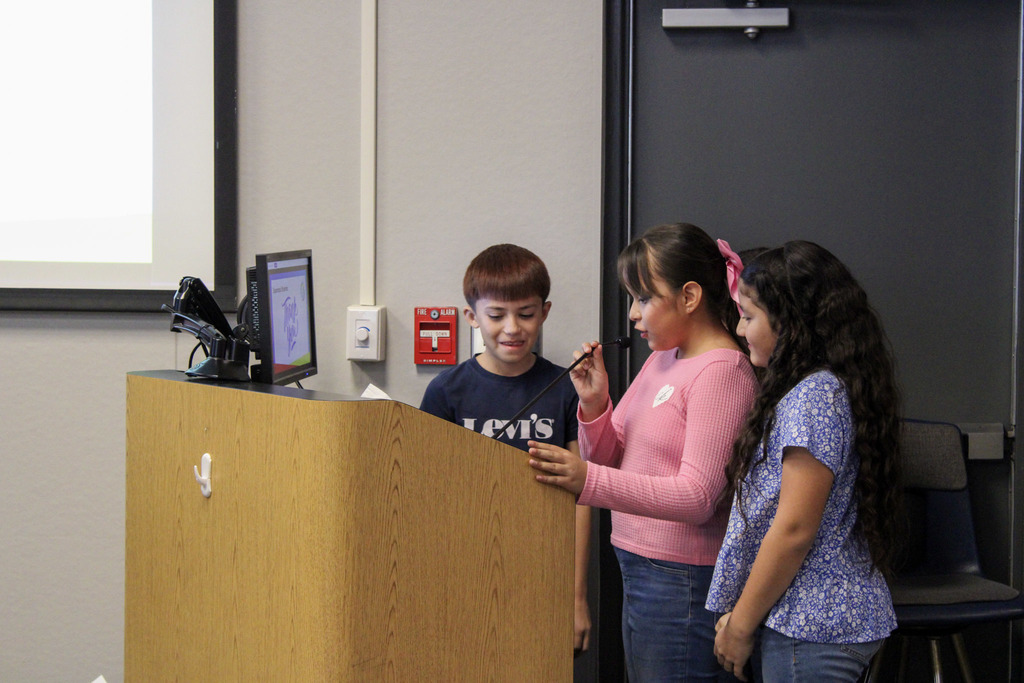 A candid shot of three of the Esperanza students standing at a wooden podium during the board meeting. One student in a pink shirt is speaking into a microphone while her peers stand beside her, looking on attentively.