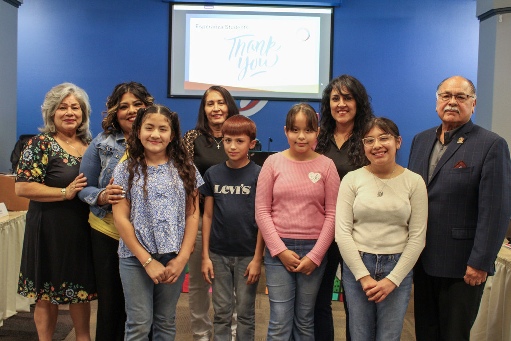 A group portrait of four elementary-aged students standing in front of four adult school board members in a meeting room. A digital screen in the background displays a "Thank You" message from "Esperanza Students". The board members and students are smiling at the camera.