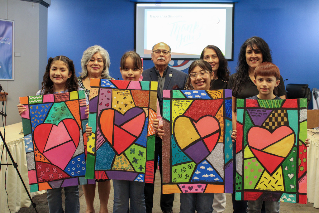The same group of students and board members, but now each student is proudly holding up one of the large, vibrant heart paintings they created. Each painting features a colorful heart divided into geometric segments with a "thank you" message at the top.