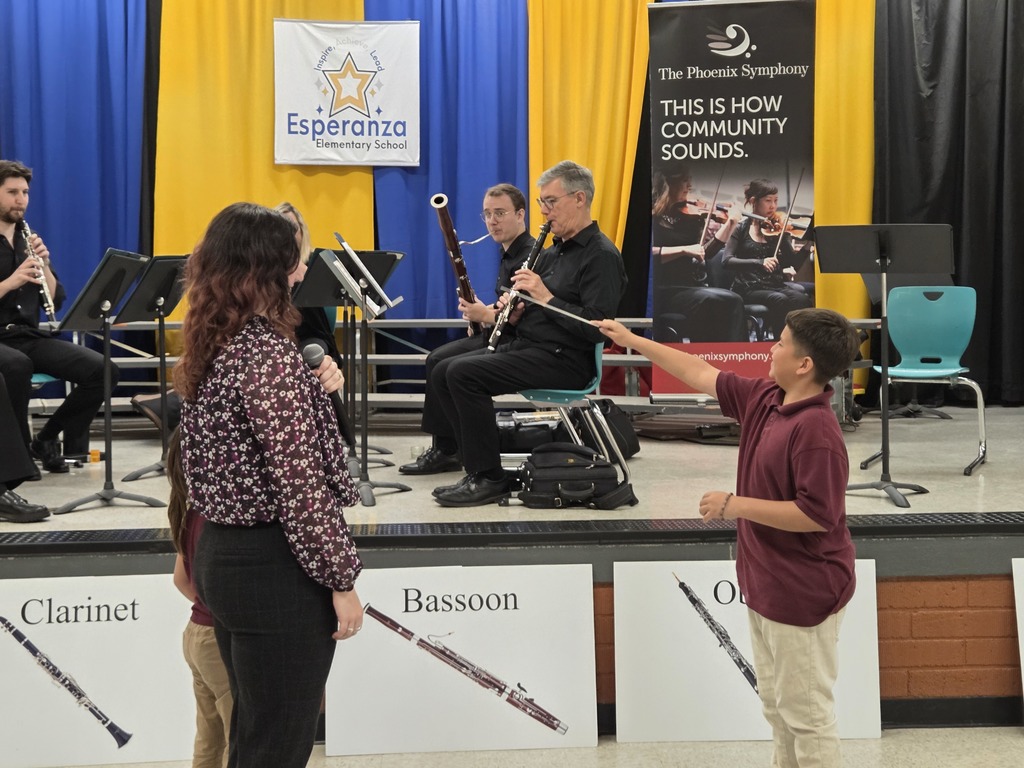A medium shot focusing on the "conducting" activity. A young boy in a maroon polo shirt stands on the floor, extending his arm toward the musicians on stage as if leading them. A symphony representative stands nearby with a microphone, smiling at the interaction. In the background, musicians are seated and playing their woodwind instruments.