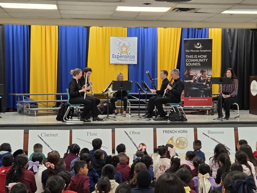 A full view of the school stage from the back of the assembly. Five musicians are seated in a semi-circle playing their instruments: a flute, oboe, French horn, bassoon, and clarinet. Below the stage, the heads of many seated students are visible, and the front of the stage is decorated with educational posters of each instrument. A banner for the Phoenix Symphony stands to the right.