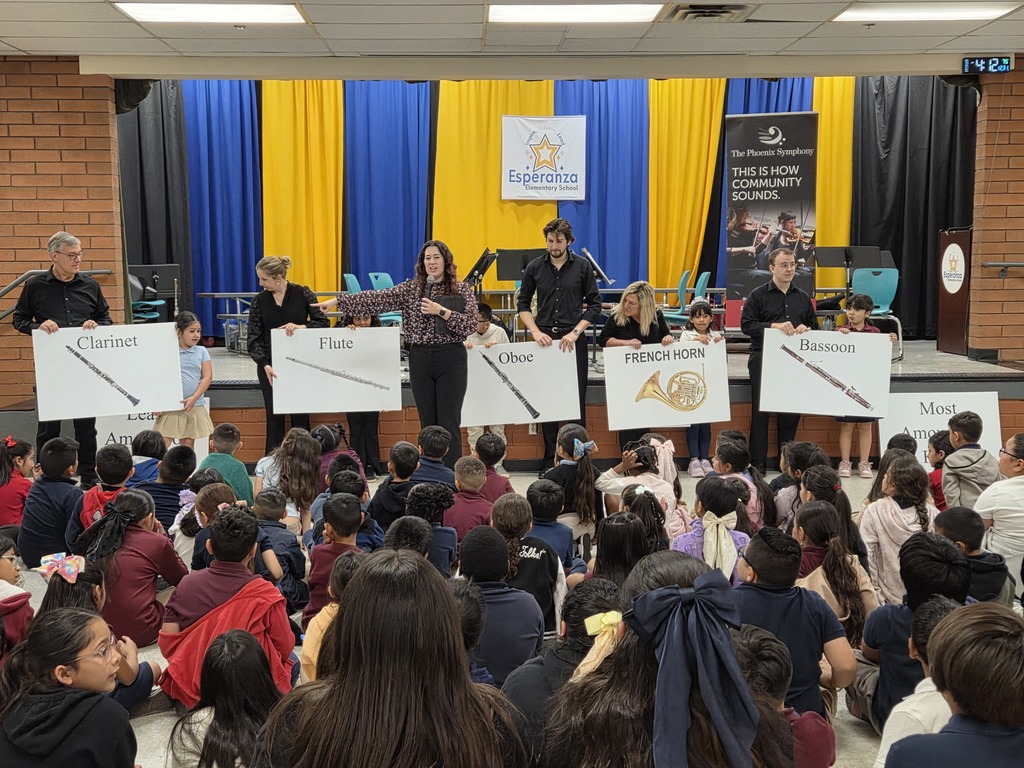 A large group of elementary students sit on the floor, facing the stage during a presentation. Five musicians and a narrator stand in a row, each holding large white signs featuring pictures and labels for the clarinet, flute, oboe, French horn, and bassoon. The narrator in the center is speaking into a microphone, pointing toward the flute player.