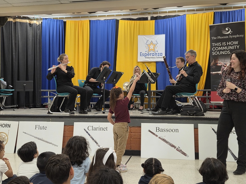 A wide shot of the Esperanza Elementary stage where a quintet from the Phoenix Symphony is performing in front of blue and yellow curtains. In the foreground, a young female student stands with her back to the camera, raising her hand high to "conduct" the musicians. Large posters identifying the flute, clarinet, and bassoon are lined up along the front of the stage.