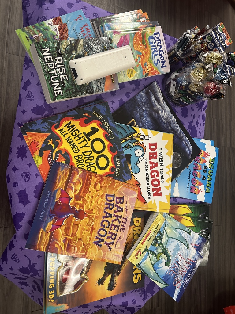 A top-down view of a table covered in a purple dragon-themed cloth. It is piled with dragon-themed books such as Wings of Fire, The Bakery Dragon, and The Rise of Neptune.
