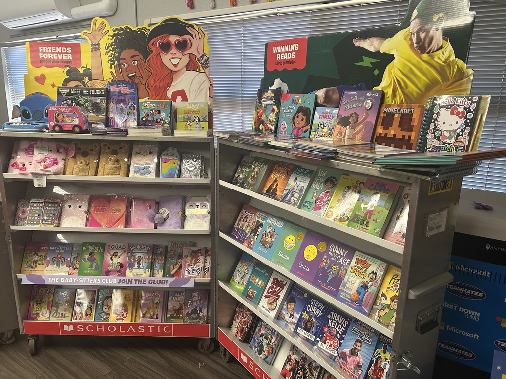A Scholastic display rack featuring "Winning Reads." Top shelves hold Bluey, Moana 2, and Minecraft books. Lower shelves feature The Baby-Sitters Club series, various journals with plush covers, and sports biographies including Steph Curry and Travis Kelce.