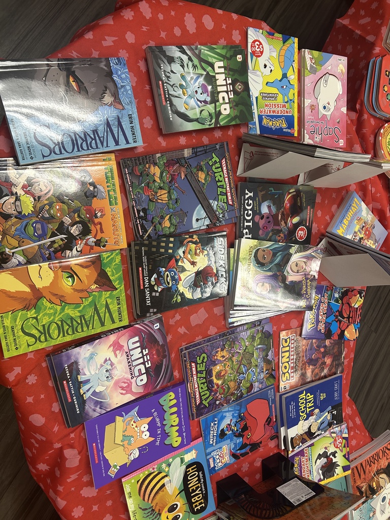 An assortment of graphic novels and manga-style books on a red cloth, including Warriors by Erin Hunter, Teenage Mutant Ninja Turtles, Sonic the Hedgehog, and Unico.