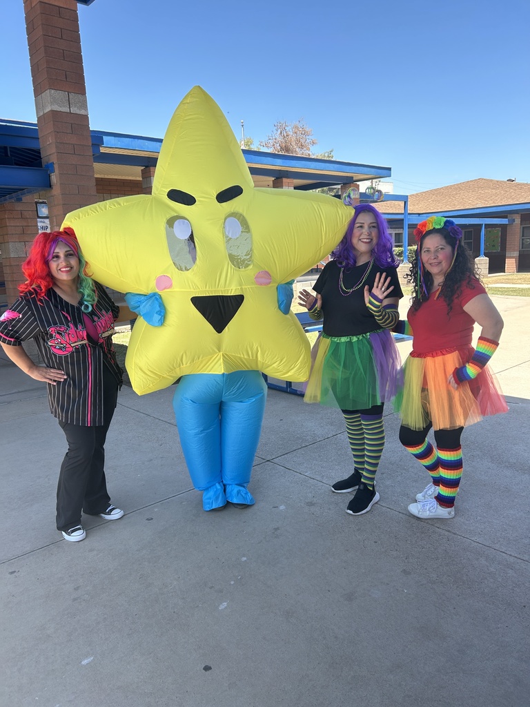 A full-length outdoor photograph taken on a paved concrete school plaza shows four people. Center stage is the large, inflatable yellow star mascot, which has small blue arms and legs, a smiling face, and pink cheeks. To the mascot’s left is a smiling woman in the red and black striped baseball jersey from image 0. To the mascot’s right are two costumed volunteers in tutus and striped leggings from image 1. They are standing under bright sunlight in front of a modern brick and blue-trimmed school building.